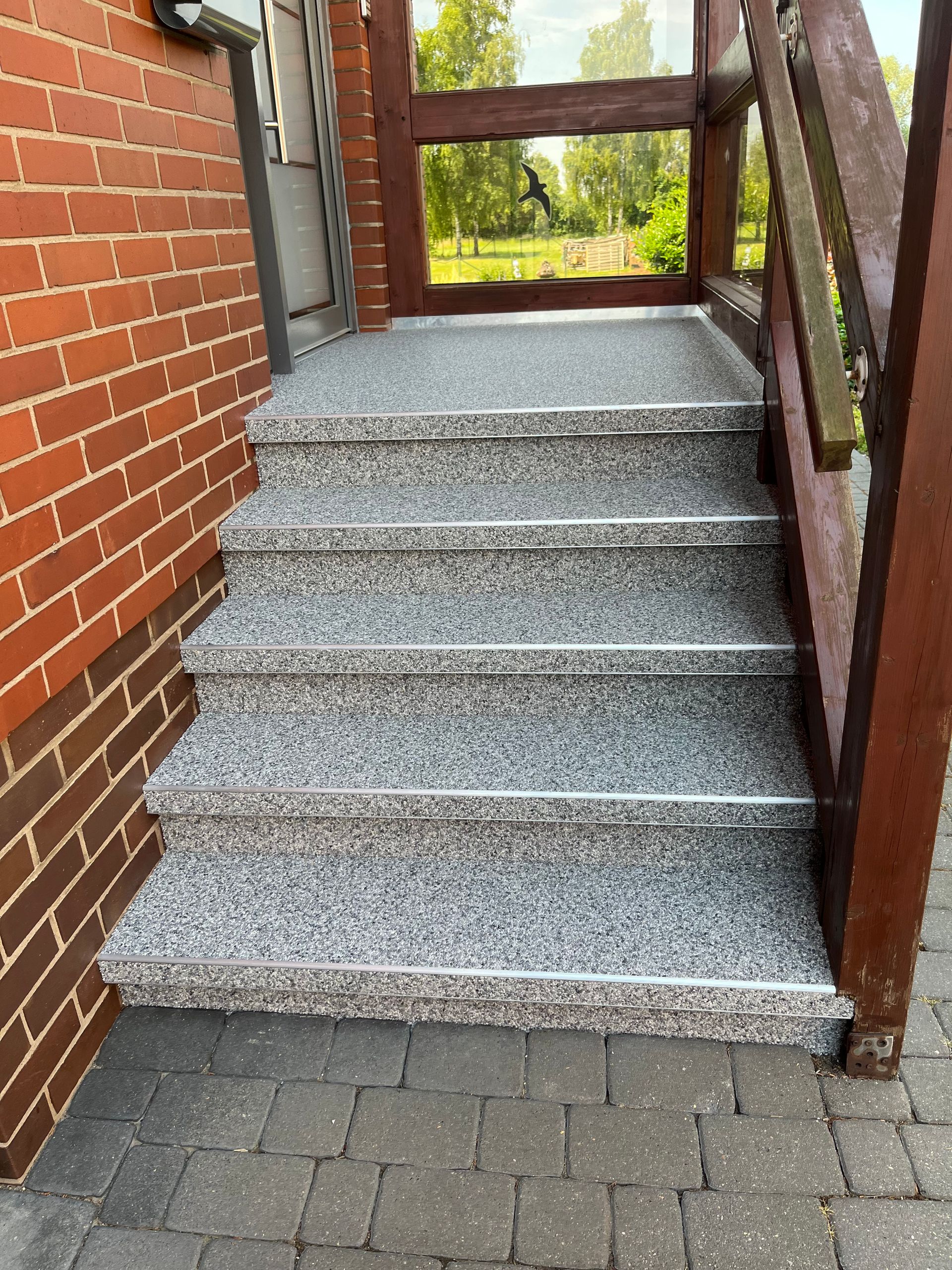 Außenansicht eines Backsteingebäudes mit grau gesprenkelten Stufen, die zu einem Holzdeck mit Geländer führen.