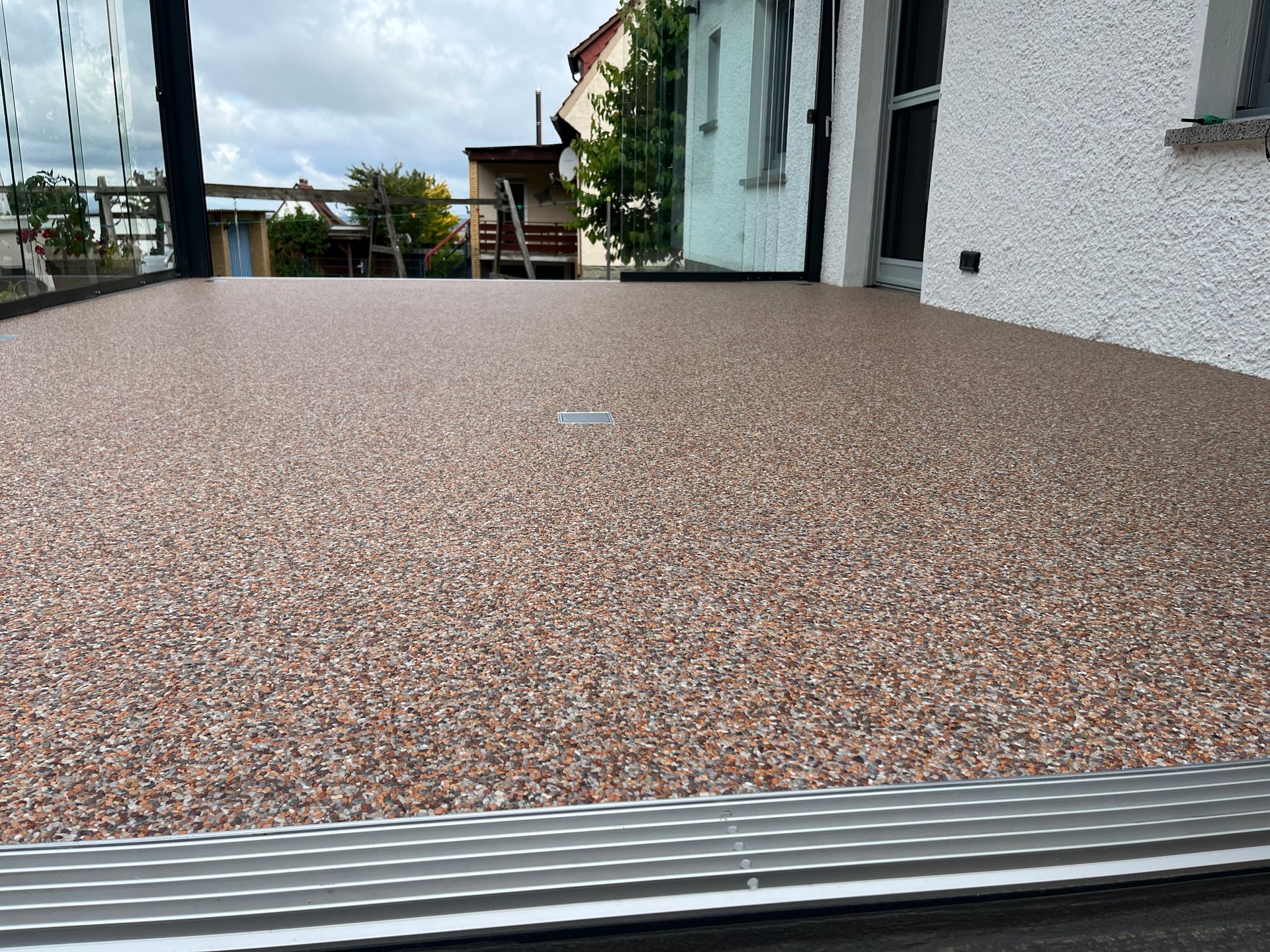 Eine flache, strukturierte Außenfläche mit kleinen, rotbraunen Steinen. Im Hintergrund sind ein Gebäude und ein Geländer zu sehen.