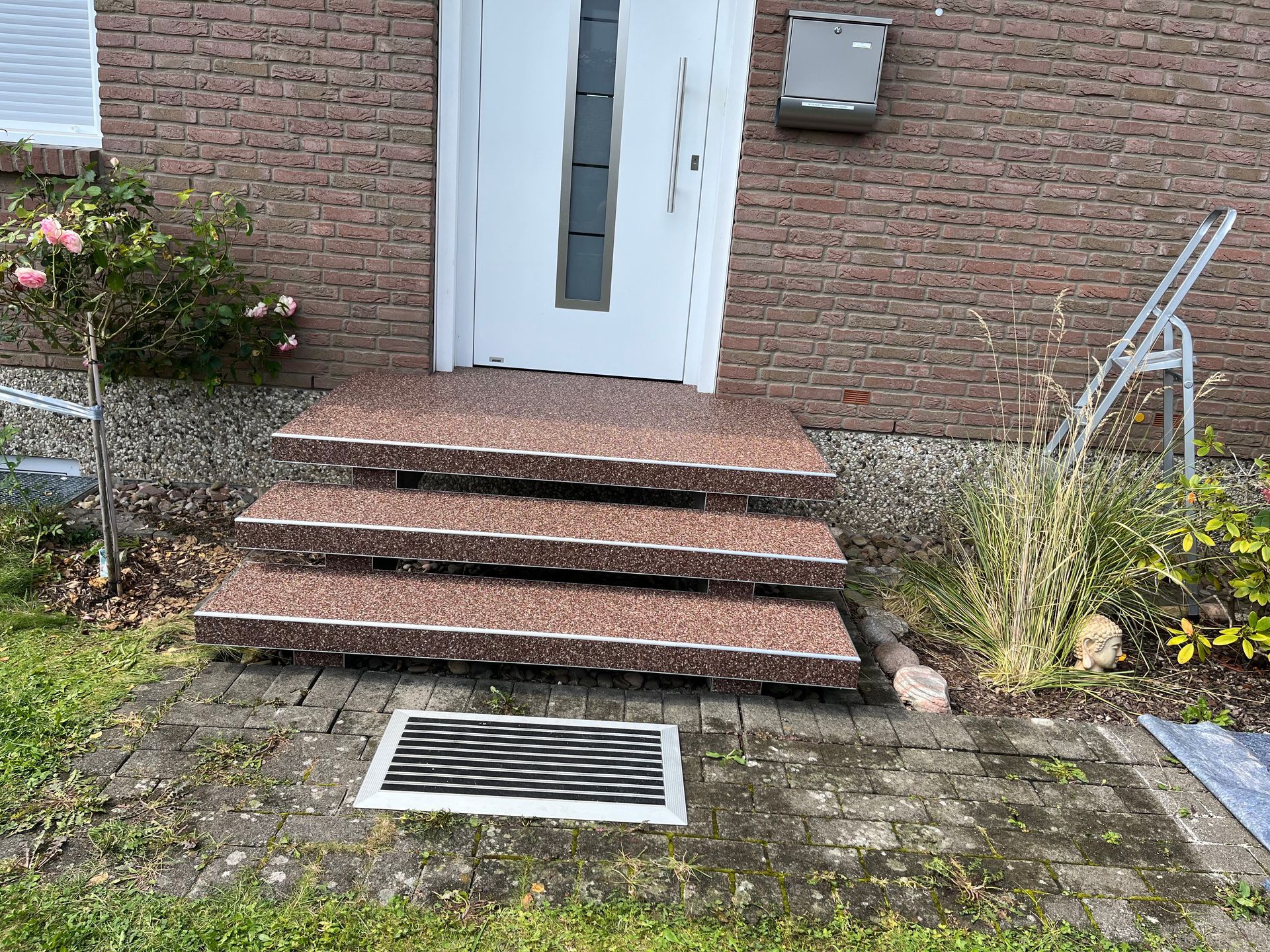 Eingang zu einem Backsteingebäude mit drei Stufen, die mit braun gesprenkeltem Material bedeckt sind. Oben befindet sich eine weiße Tür und unten ein Metallgitter.