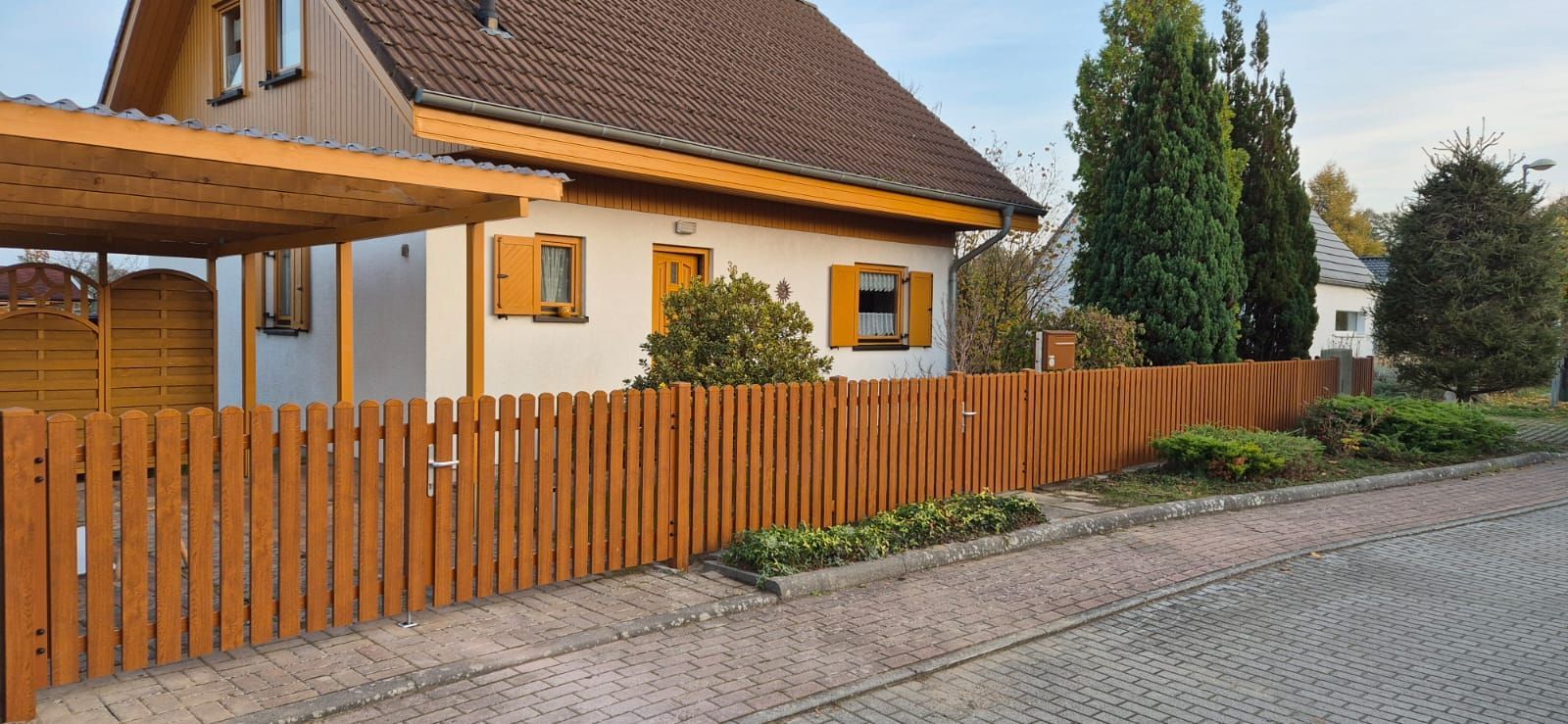 Ein brauner Holzzaun umgibt ein Haus mit braunem Dach und gelben Fensterläden. Das Haus liegt an einer gepflasterten Straße und verfügt über einen Carport.