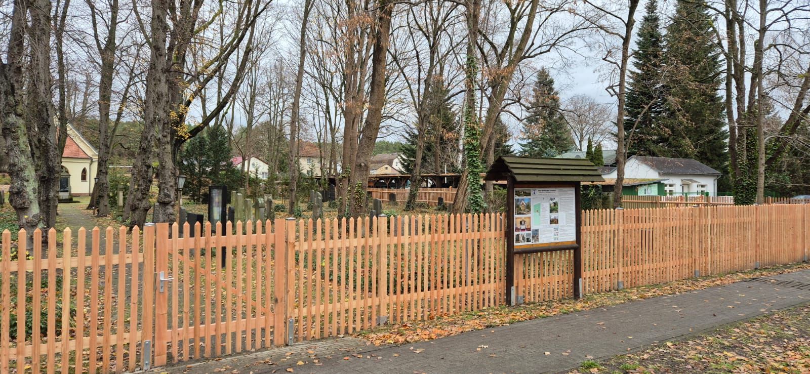 Ein Holzzaun mit einem Tor umgibt ein Grundstück mit Gebäuden und Bäumen, und in der Nähe des Zauns steht ein Schild.