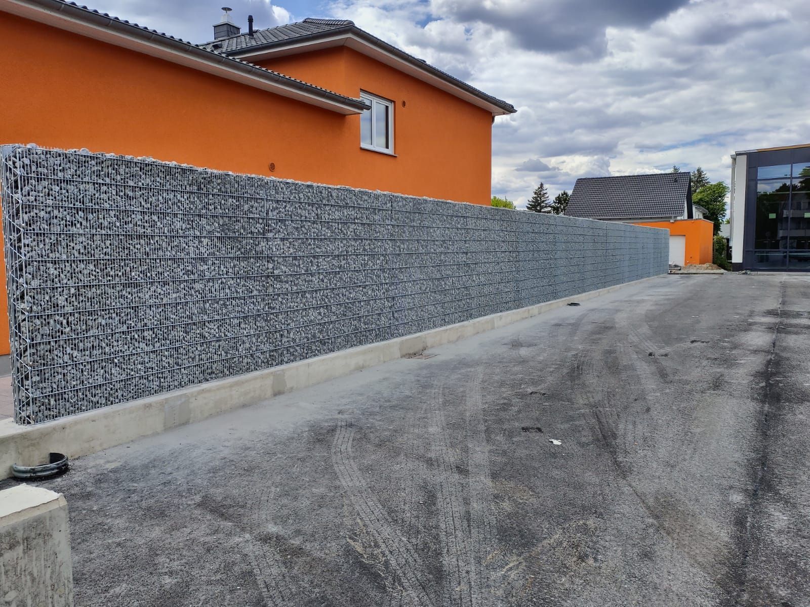 Eine lange, mit Steinen gefüllte Gabionenmauer verläuft entlang einer Kiesauffahrt vor einem orangefarbenen Gebäude.