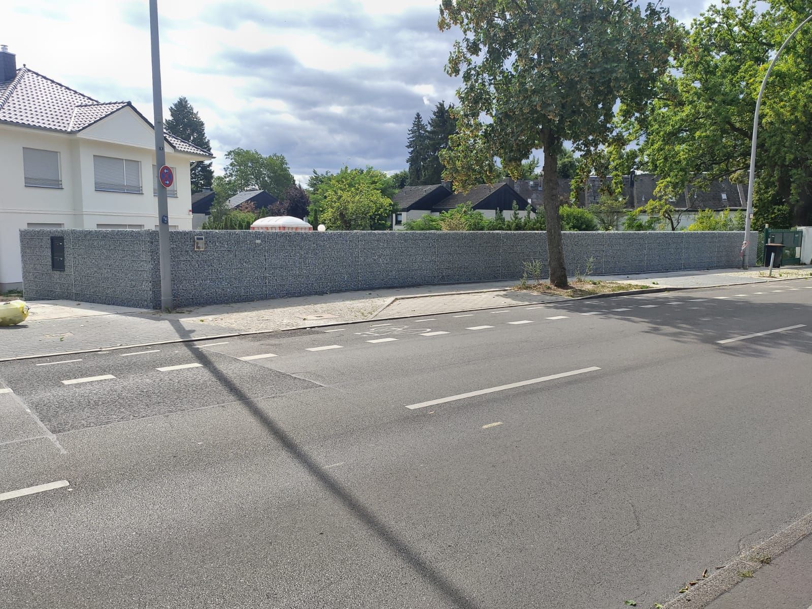 Eine lange Stützmauer aus grauem Stein säumt eine Straße neben einem Bürgersteig und einer Straße. Links davon befindet sich ein zweistöckiges weißes Gebäude.