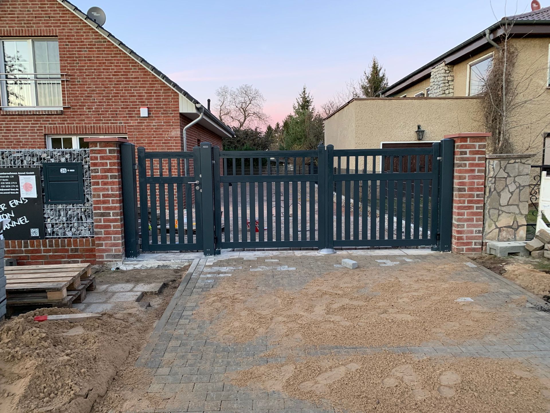 Dunkelgraues Metalltor mit vertikalen Lamellen zwischen Ziegel- und Steinpfeilern. Der Boden davor ist mit Sand bedeckt.