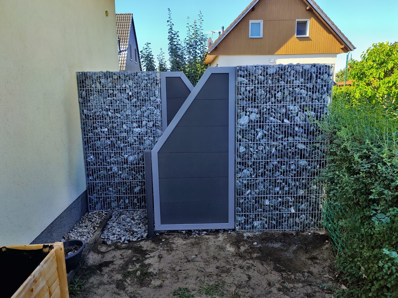 Ein grauer Gabionenzaun aus Stein mit einem dunkelgrauen Tor in einem Hof.