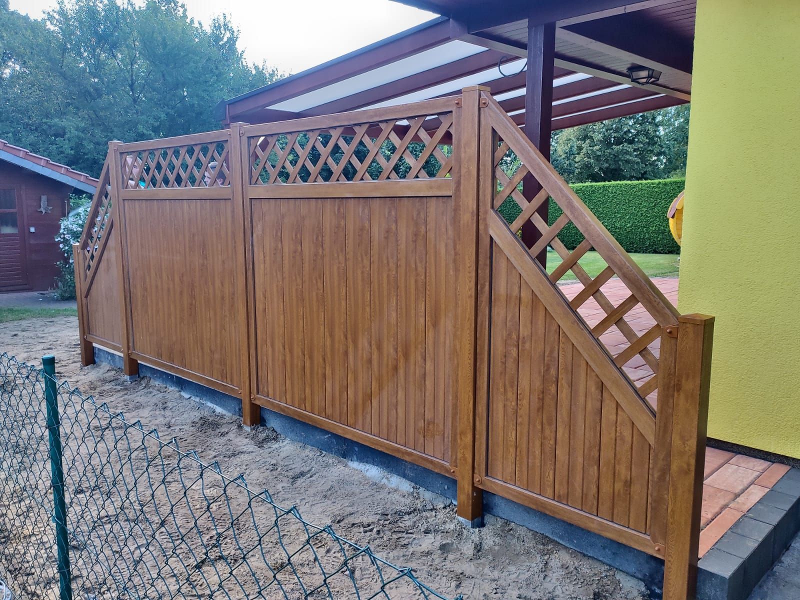 Brauner Holzzaun mit Rautengitter-Oberteil, vor einem Haus und einem Carport, auf einem Kiesboden.