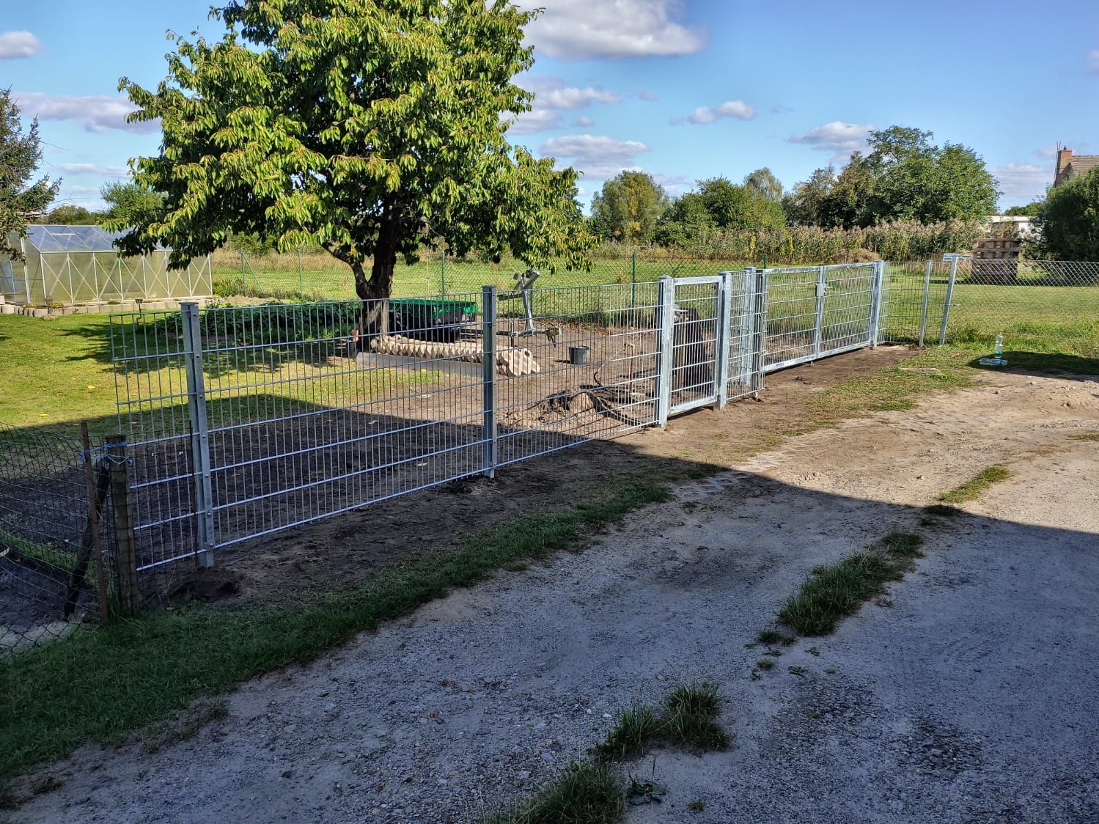 An einem sonnigen Tag umgibt ein Metallzaun teilweise ein Gartengrundstück. Ein Feldweg führt zum Tor.