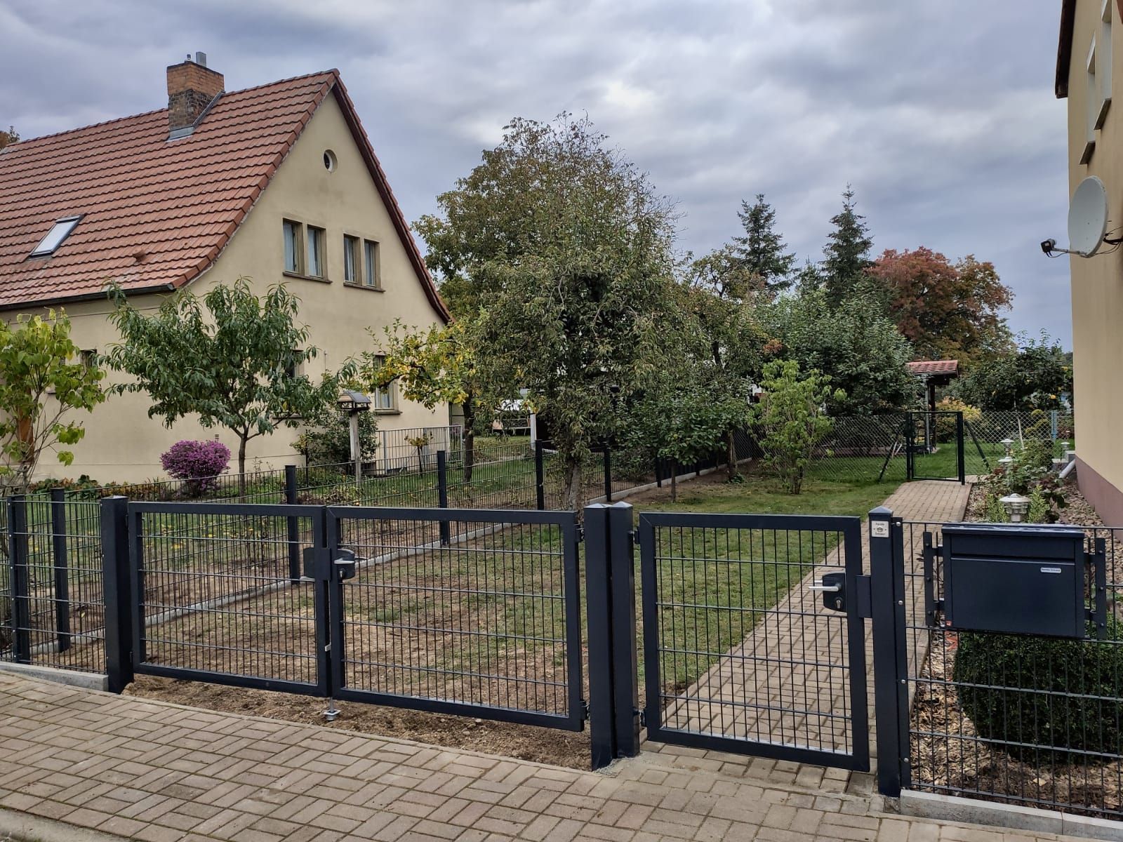 Ein Wohngrundstück mit schwarzem Metallzaun, Tor und Briefkasten. Zu sehen ist ein Haus mit rotem Ziegeldach und einem Garten mit Bäumen.