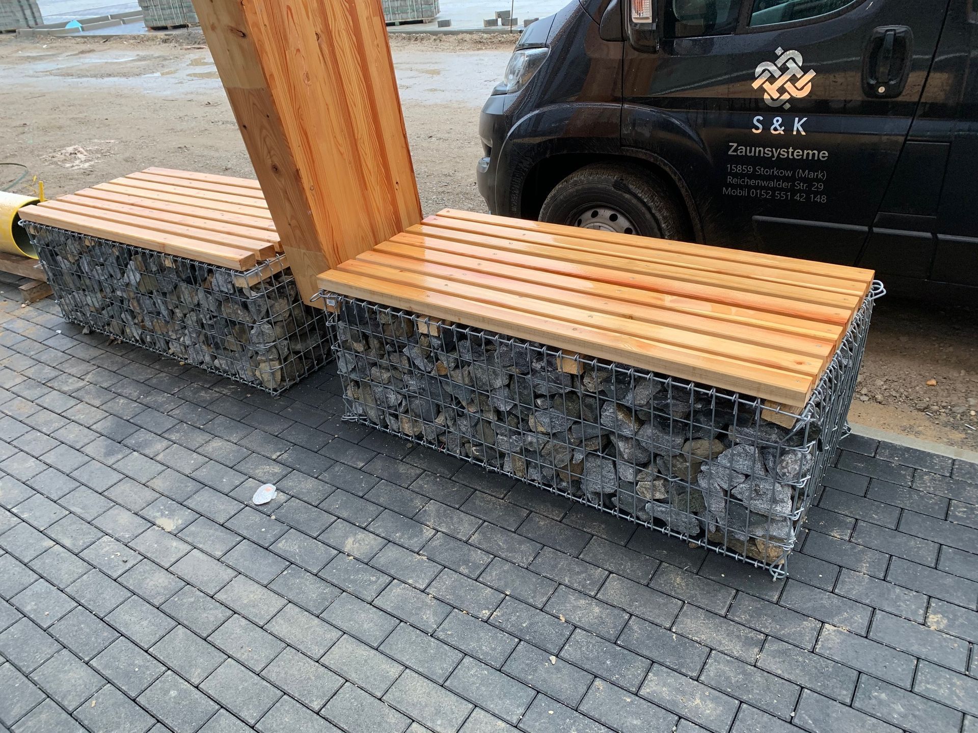 Zwei Außenbänke mit Steinsockeln und Holzsitzen neben einem schwarzen Lieferwagen auf einer gemauerten Terrasse.