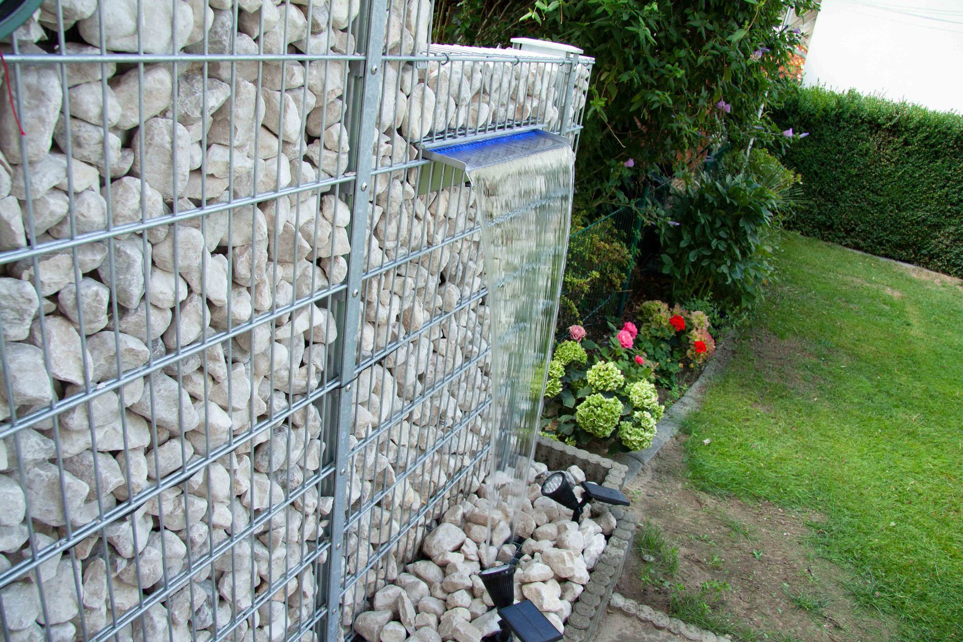 Gabionenwand mit Wasserfall; weiße Steine in Metallfassung, Wasser fließt auf verstreute Felsen. Grüne Pflanzen und Gras umgeben die Wand.