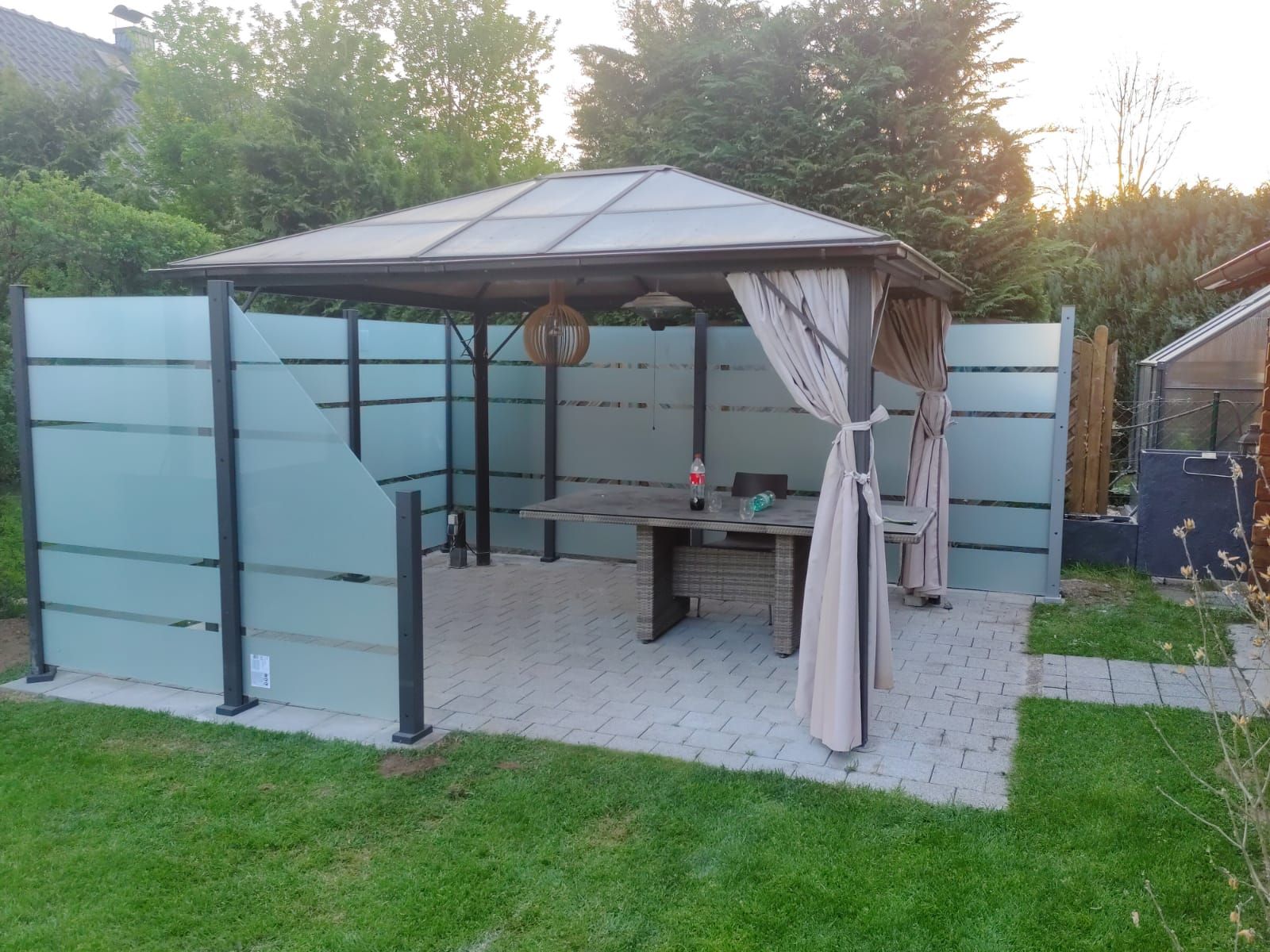 Auf einer gepflasterten Terrasse steht ein Pavillon mit Milchglaswänden und beigen Vorhängen mit einem Tisch, umgeben von einer Rasenfläche.