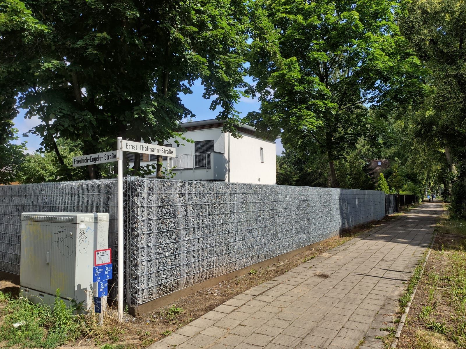 Eine lange Gabionenmauer umgibt ein Gebäude entlang eines Gehwegs. Links sind ein Straßenschild und ein Versorgungskasten zu sehen. Im Hintergrund sind Bäume zu sehen.