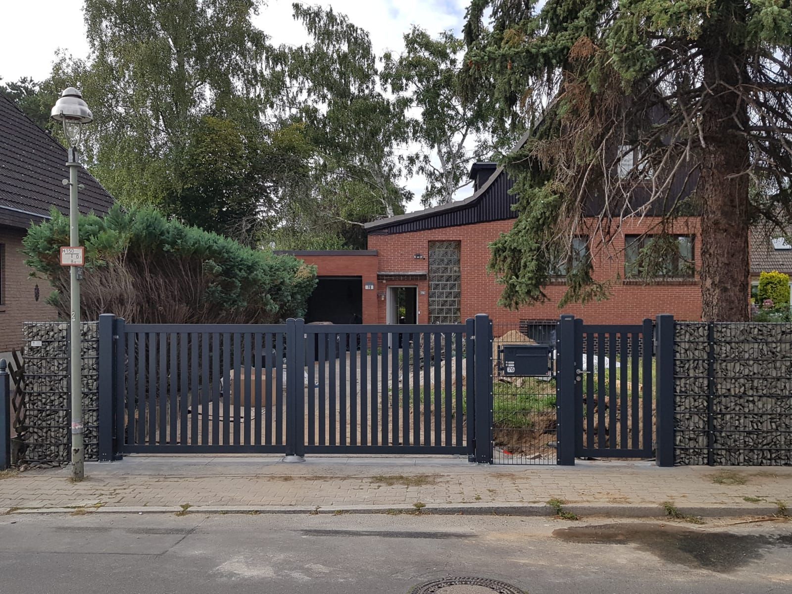 Ein dunkelgraues Metalltor und ein Zaun umschließen ein rotes Backsteinhaus mit Bäumen im Hintergrund. Im Vordergrund die Straße.