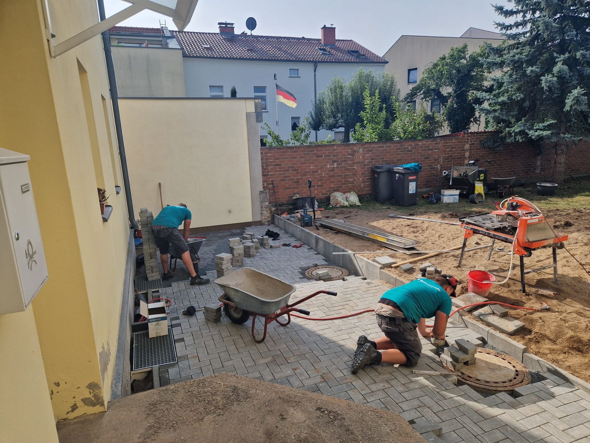 Zwei Arbeiter verlegen Pflastersteine in einem Hinterhof. Dabei sind eine Schubkarre und ein Betonmischer im Einsatz. Im Hintergrund weht eine deutsche Flagge.