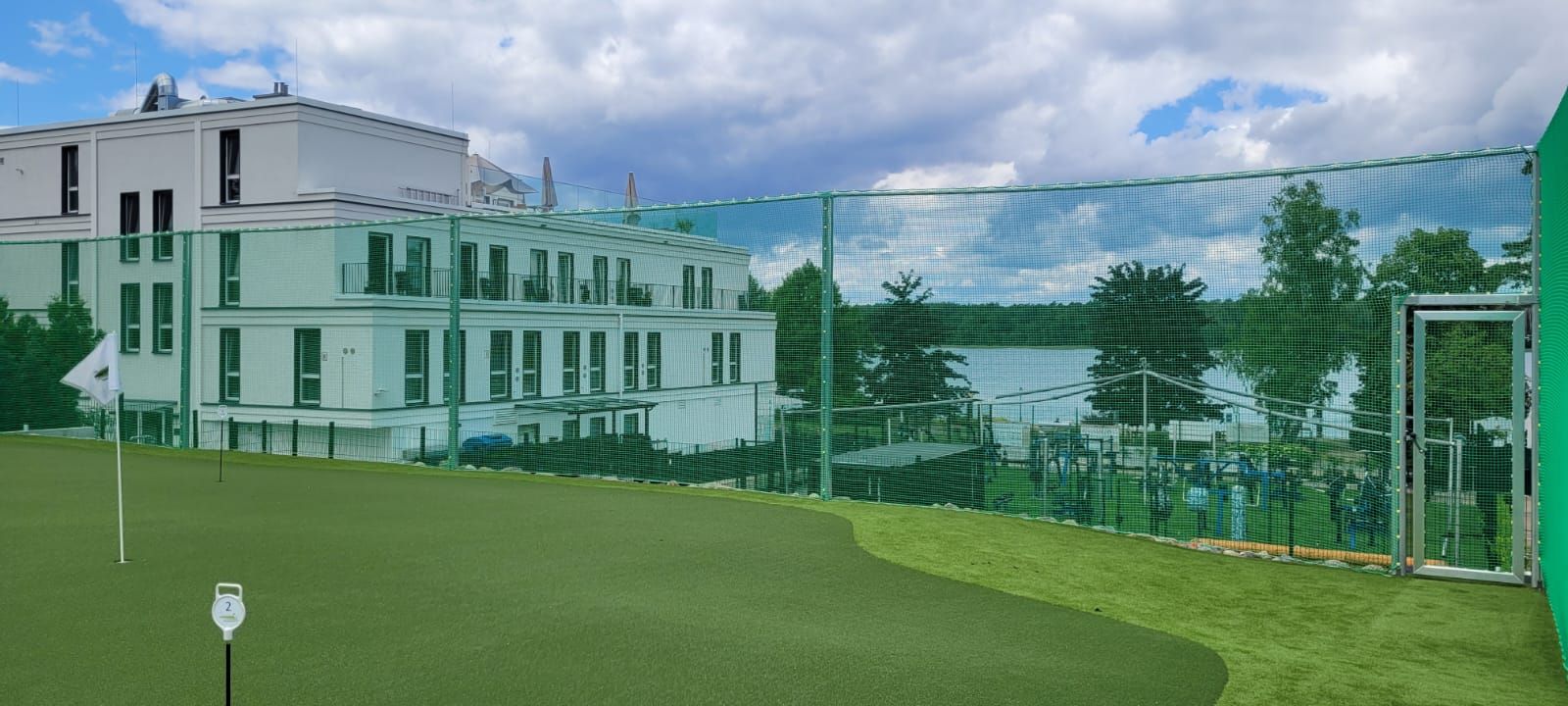 Ein Golfplatz mit einem weißen Gebäude, einem See und einem grünen Netz. Der Himmel ist bewölkt und das Gras ist grün.
