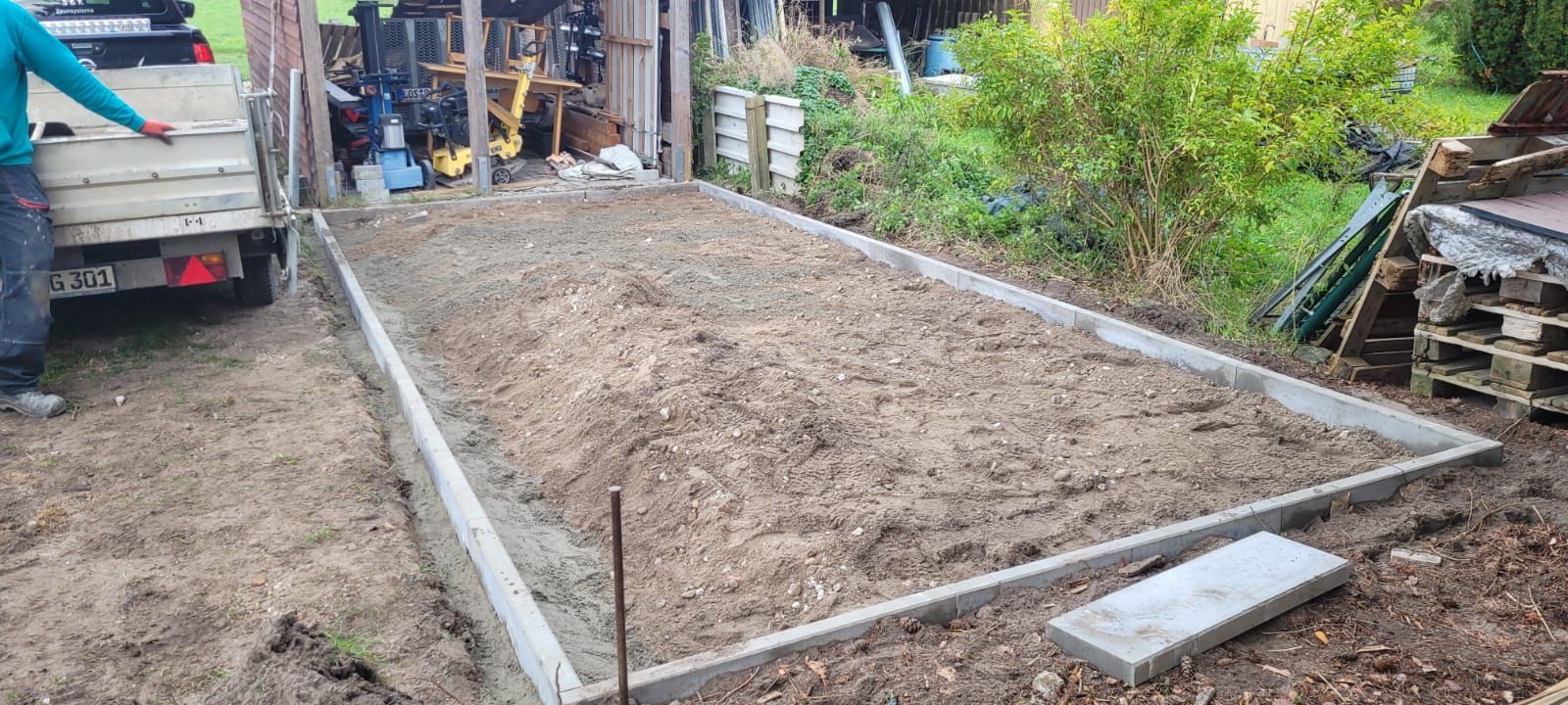 Ein rechteckiges Gartenbeet mit Betonrändern ist mit lockerer Erde gefüllt. Eine Person steht neben einem Anhänger, der neben dem Beet geparkt ist.
