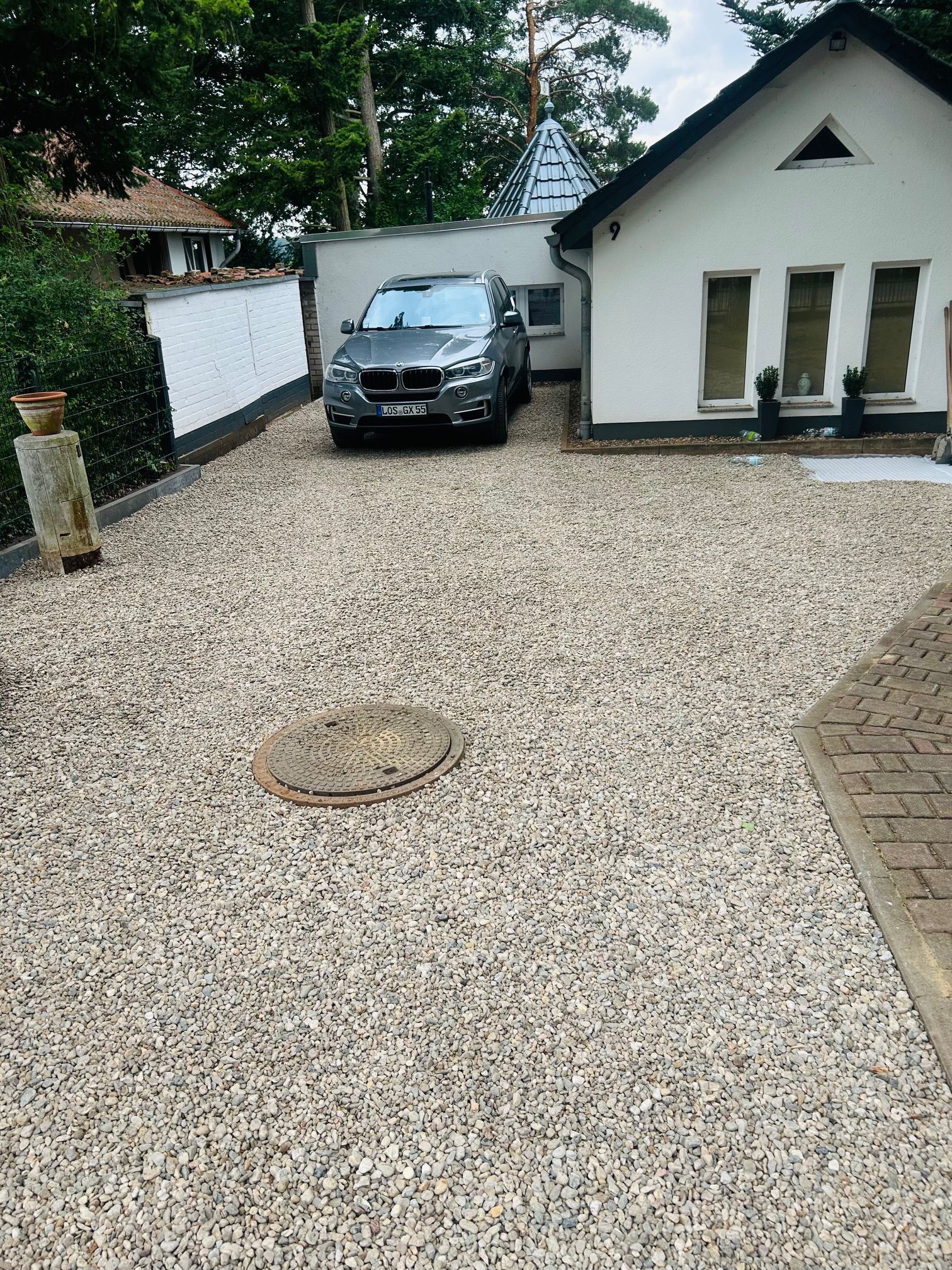 Eine Kiesauffahrt mit einem vor einem weißen Haus geparkten Auto. Im Vordergrund ist ein Gullydeckel zu sehen.