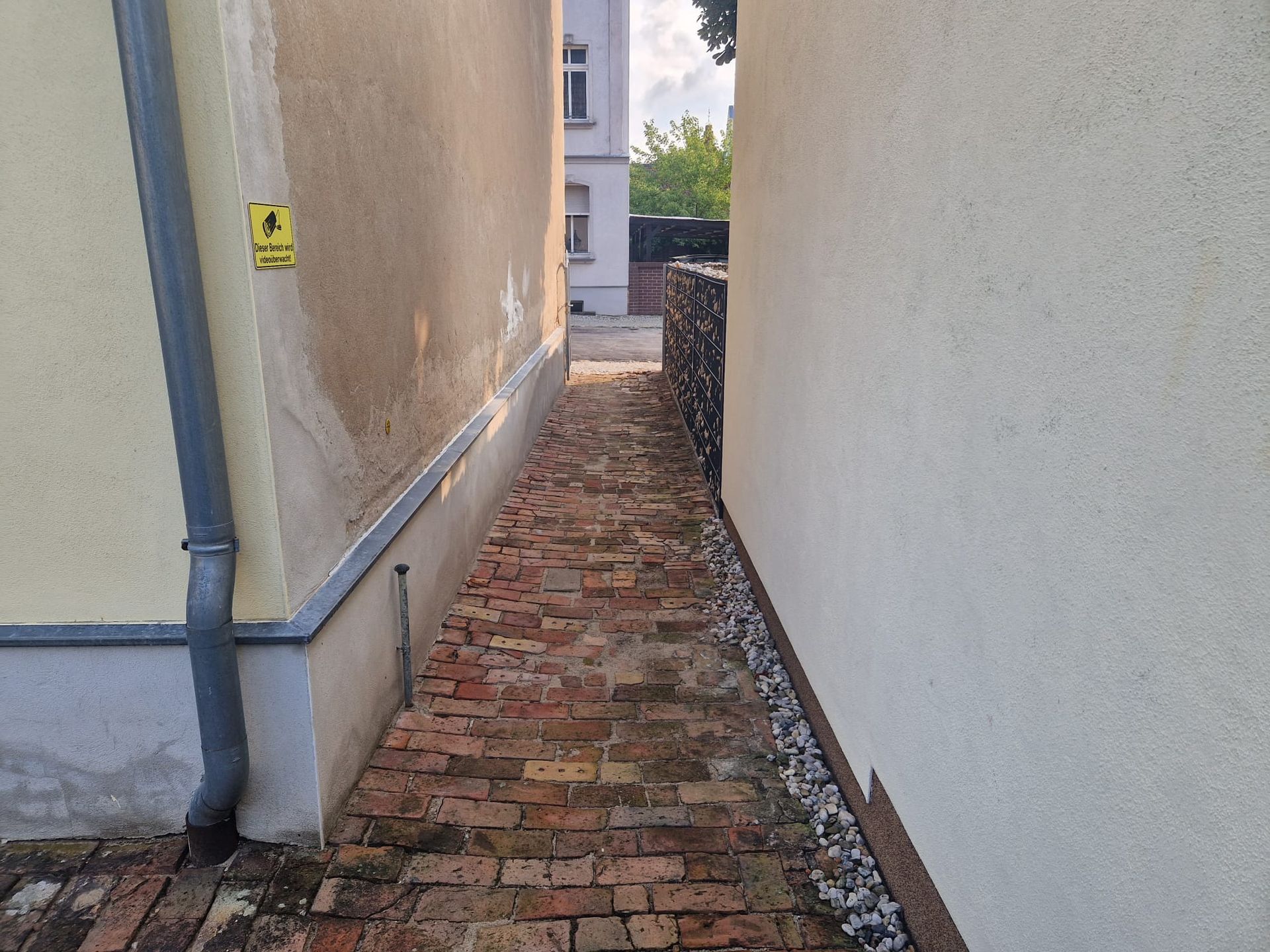 Schmale Backsteingasse zwischen zwei hellen Gebäuden, mit einer niedrigen Mauer auf der rechten Seite und dem sichtbaren Himmel am Ende.
