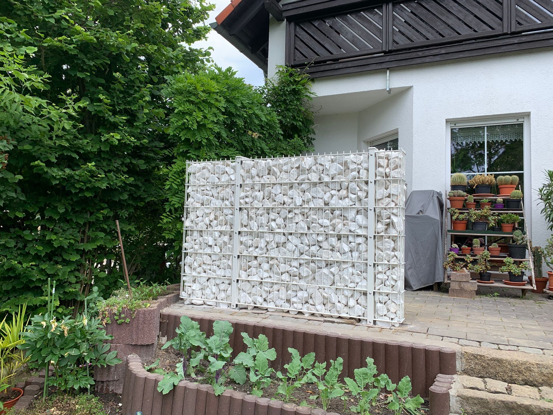 Eine weiße Gabionenwand voller Steine steht neben einem Haus mit Pflanzen und einem Garten.