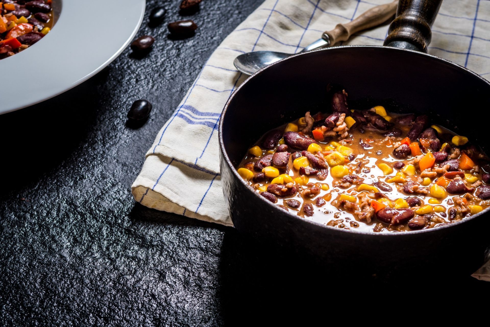 Chile en olla con maíz y frijoles, junto a un recipiente y una cuchara sobre un mantel a cuadros.