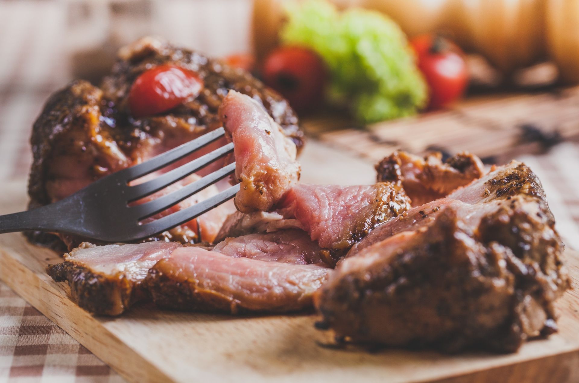 Carne asada cortada en rodajas sobre una tabla de madera, con un tenedor sujetando un trozo. Verduras al fondo.