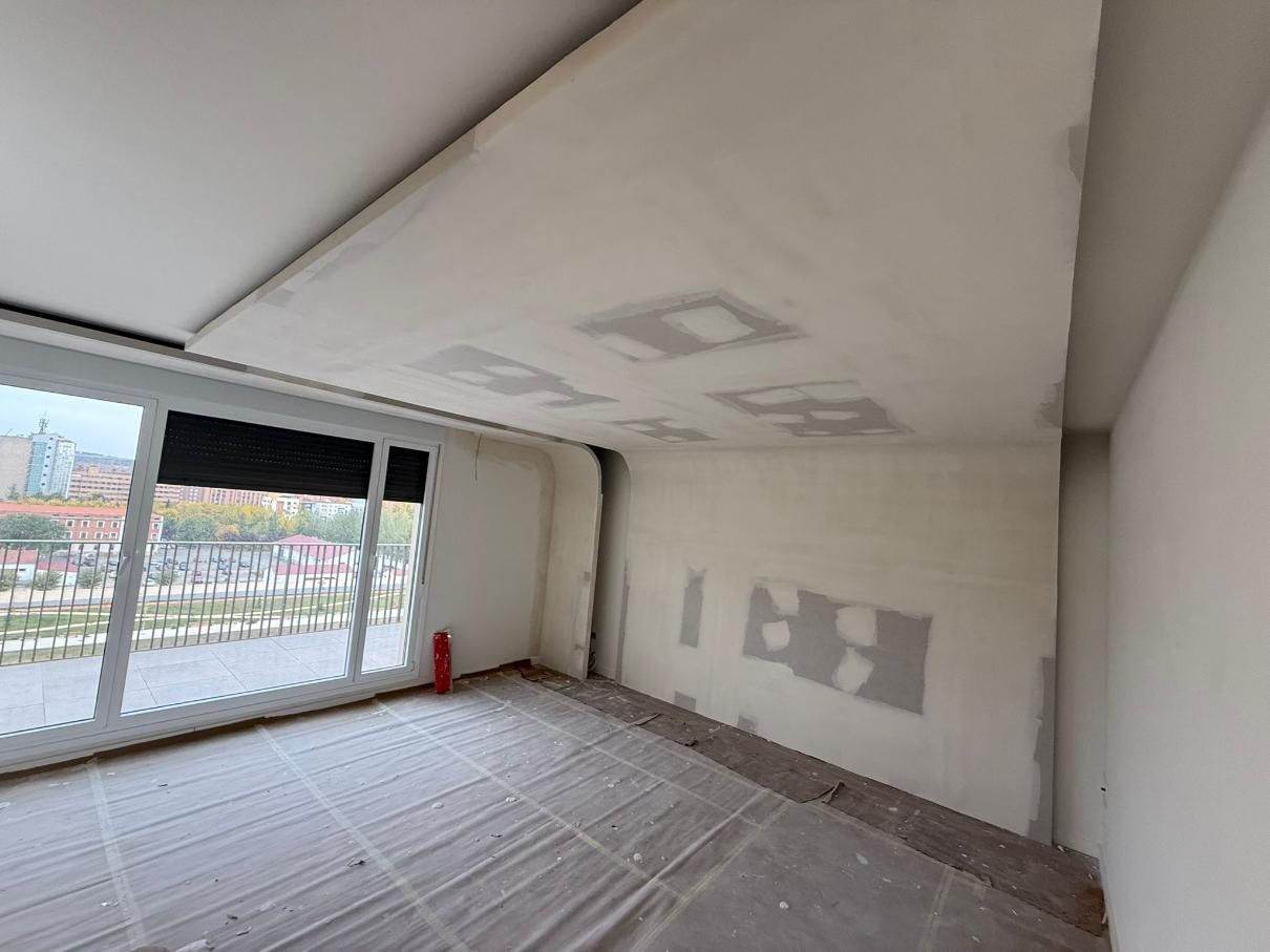 Habitación vacía con ventana grande, paneles de yeso sin terminar en paredes y techo. Plástico protector cubre el suelo.