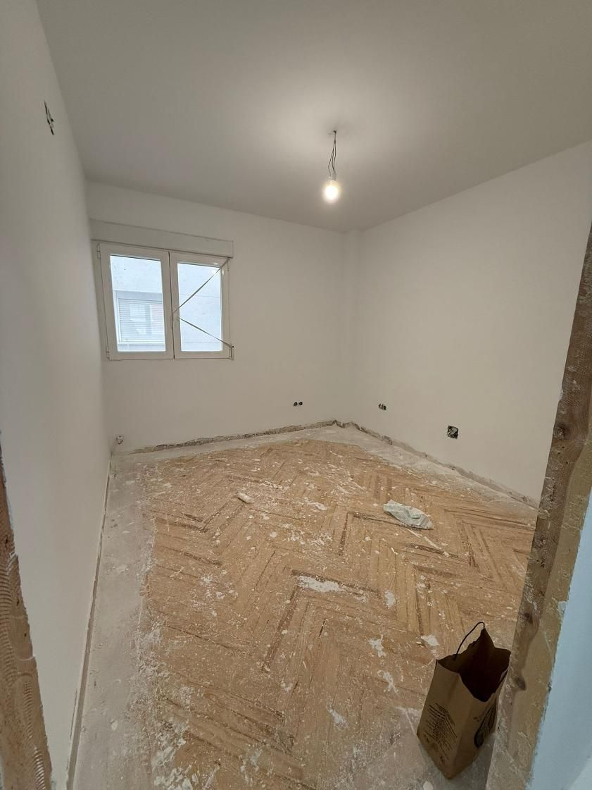 Habitación vacía con suelo de parquet cubierto de escombros, ventana y una única bombilla colgando del techo.