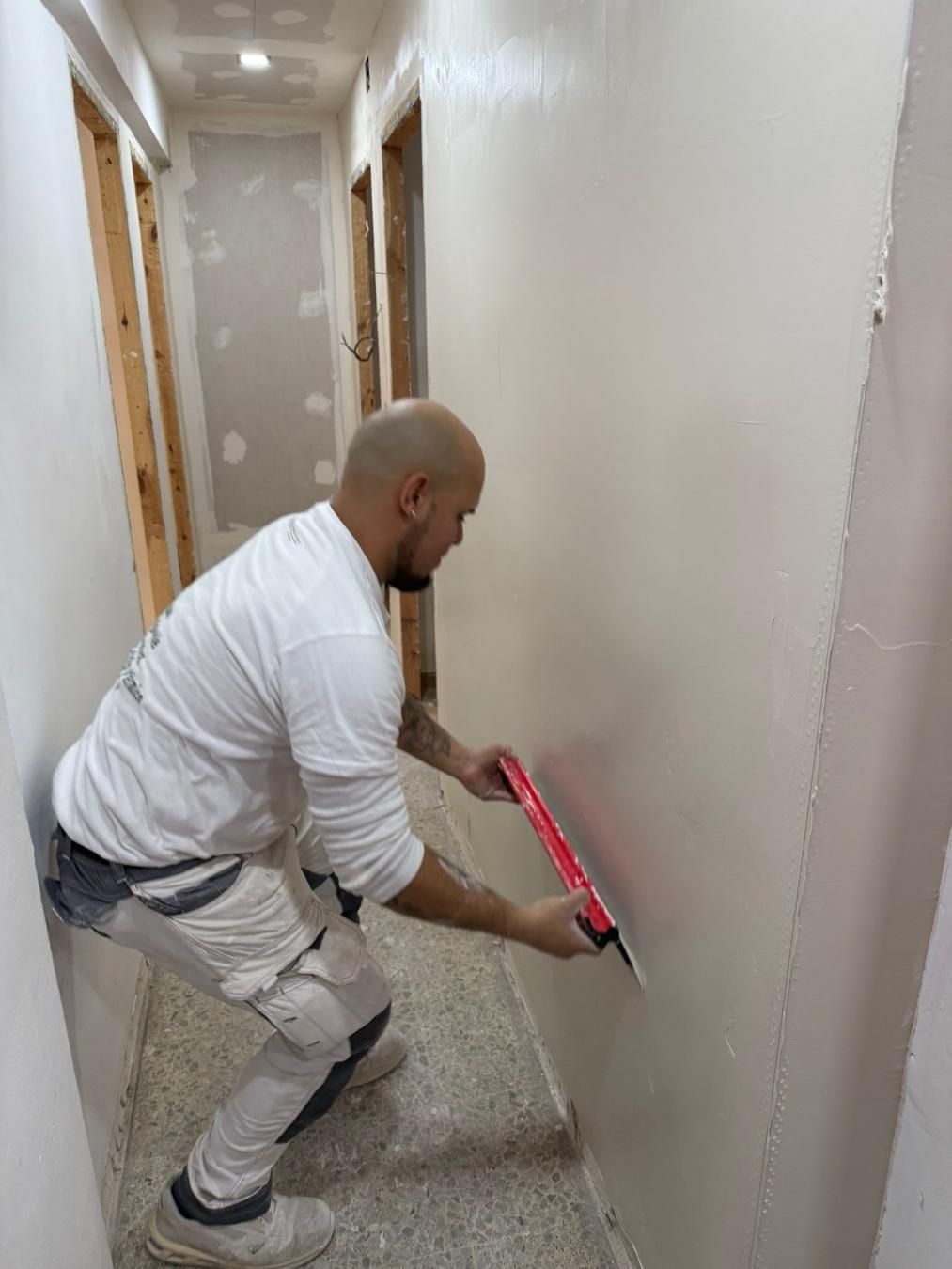 Persona alisando paneles de yeso en un pasillo con una espátula roja y plateada; paredes de color beige