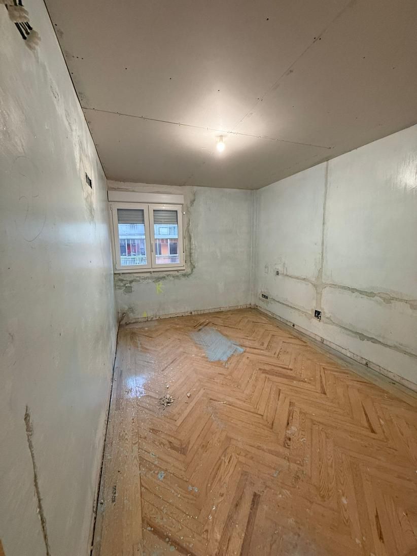 Habitación pequeña y vacía con suelo de parquet, ventana y paredes sin terminar.