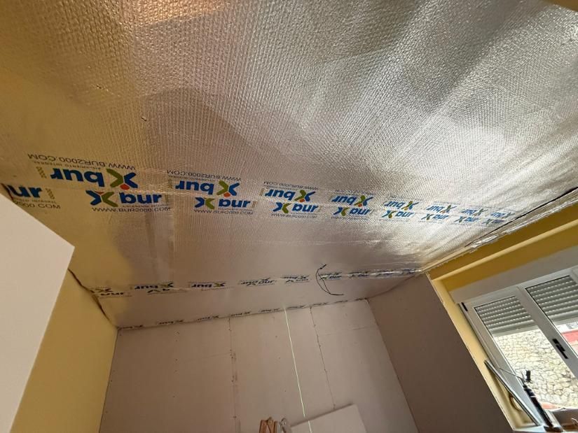 Aislamiento de techo con lámina de plata, costuras selladas, habitación con paredes amarillas, ventana.