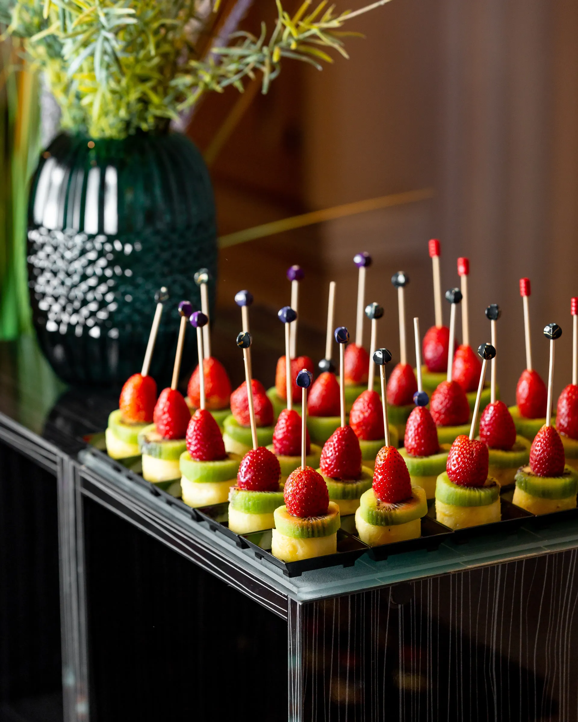 Brochetas de frutas: fresas, kiwi y piña en una bandeja negra, con un jarrón verde de fondo.
