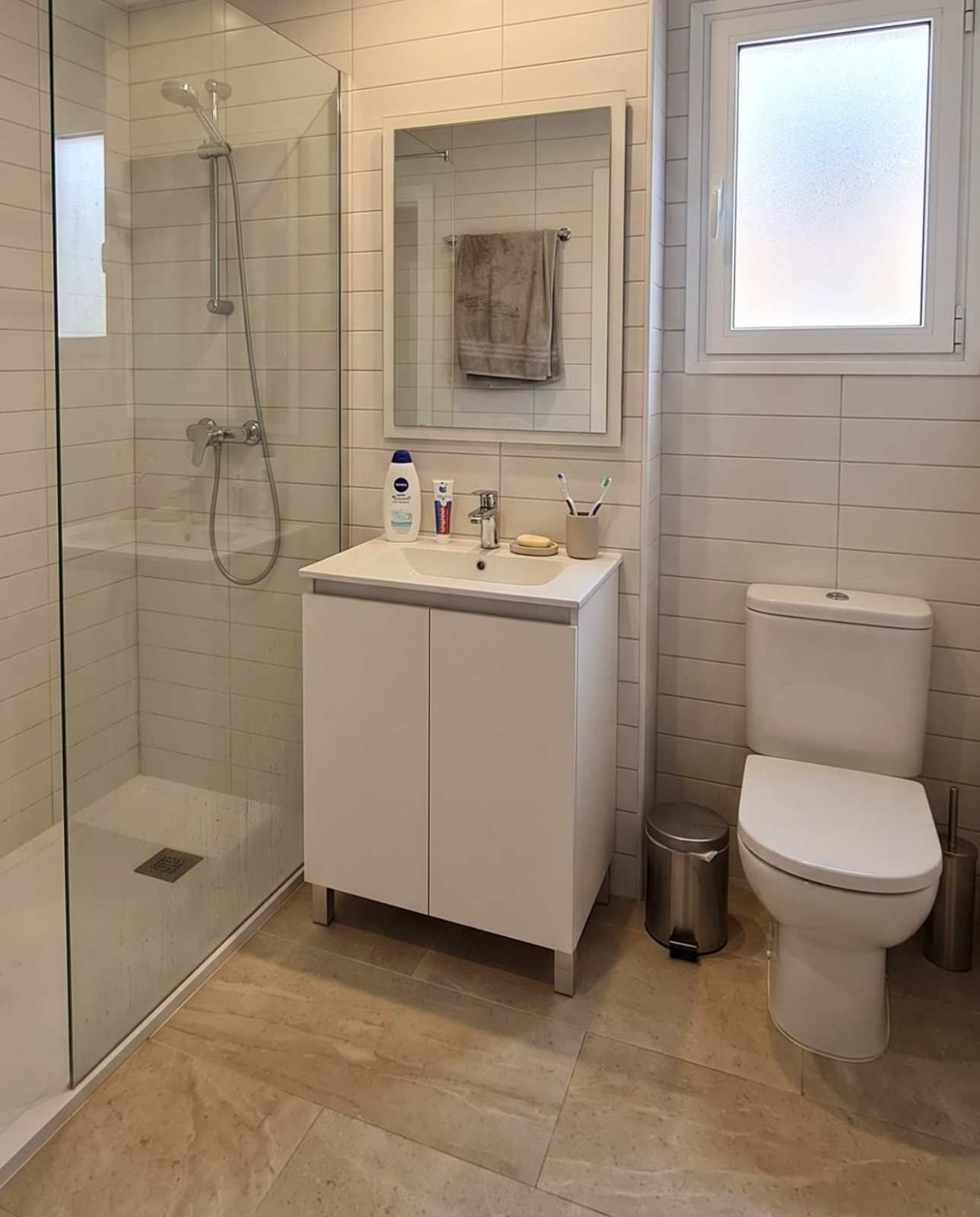 Baño moderno con ducha de cristal, mueble de lavabo blanco, inodoro, ventana y suelo de baldosas beige
