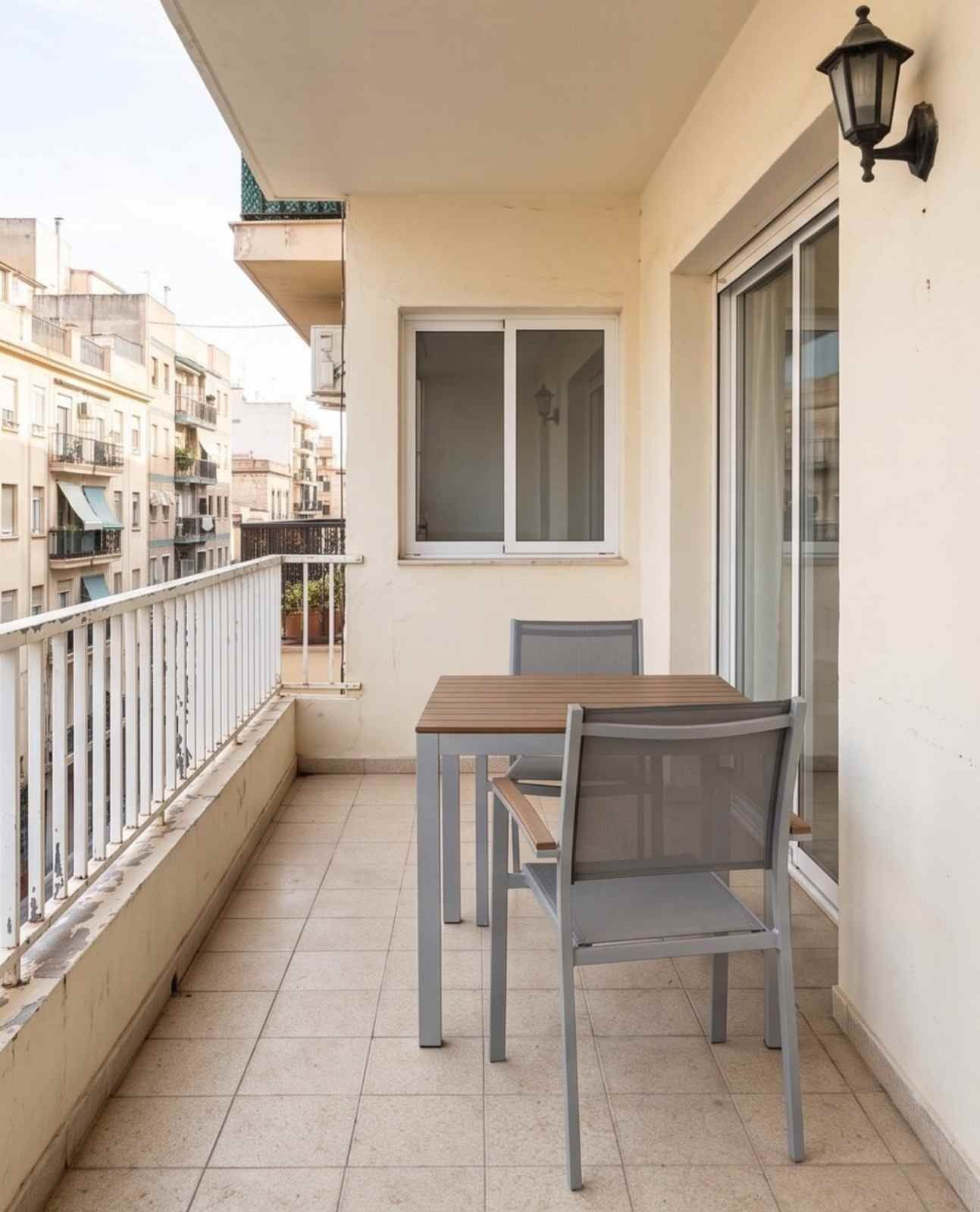 Pequeño balcón beige con una mesa, dos sillas, suelo de baldosas, barandilla y puerta corredera de cristal.