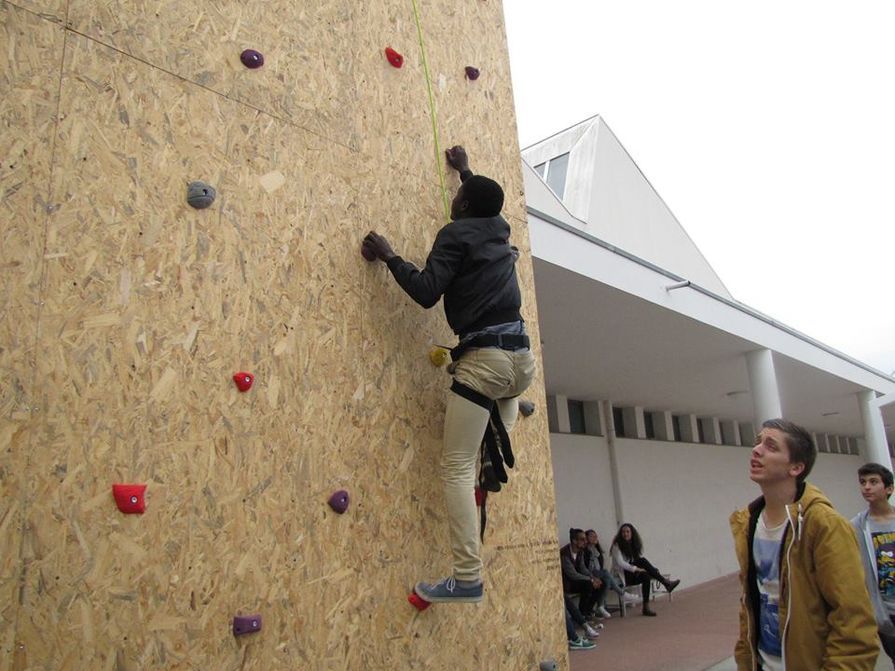 Pessoa escalando uma parede de escalada ao ar livre com espectadores ao fundo.