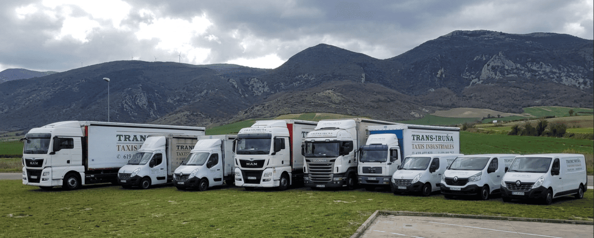 Una hilera de furgonetas blancas y camiones de reparto aparcados en un campo de hierba con montañas al fondo.