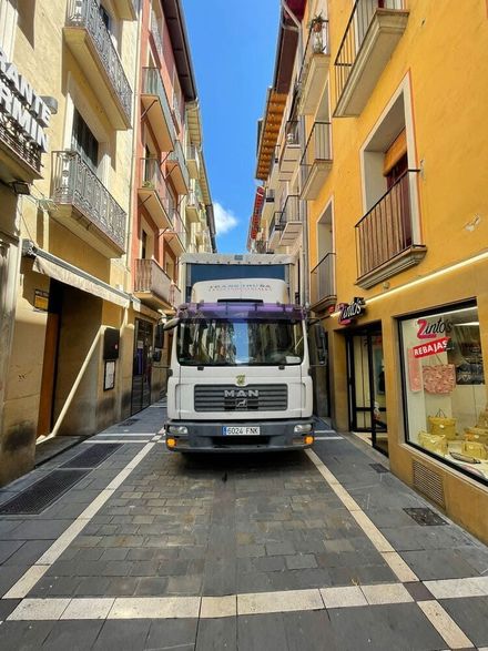 Camión blanco en una estrecha calle empedrada entre edificios amarillos bajo un cielo azul.