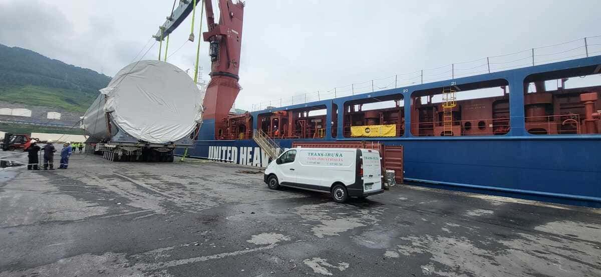 Un buque de carga descarga un gran tanque cilíndrico blanco en un puerto, con una grúa y una furgoneta cerca.