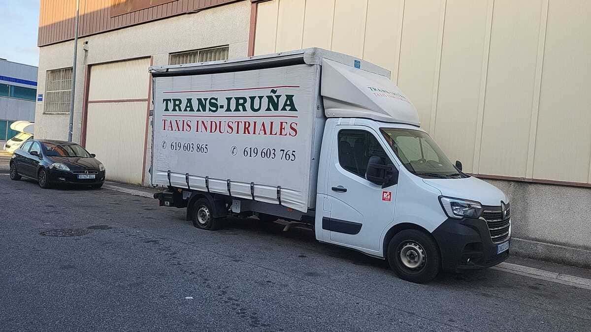Camión de reparto blanco con el logotipo “TRANS-IRUNA” estacionado junto a la pared de un almacén en un terreno de grava.