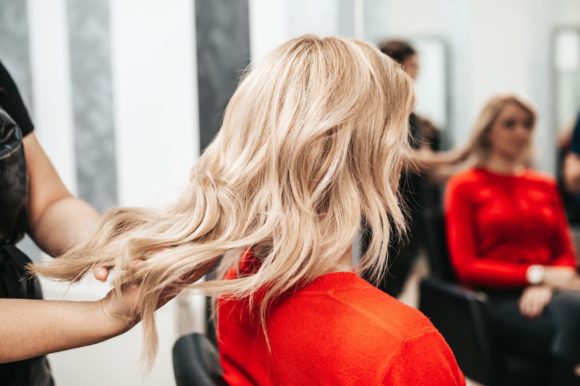 Une femme en chemise rouge a les cheveux blonds coiffés par un coiffeur dans un salon.