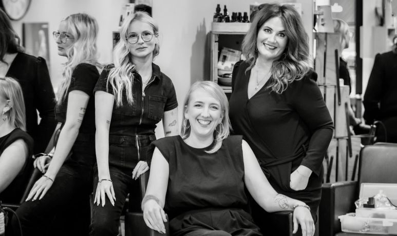 Groupe de cinq femmes souriantes dans un salon de coiffure. La plupart sont vêtues de noir.