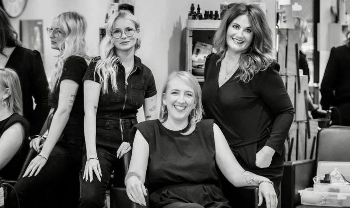 Groupe de cinq femmes souriantes dans un salon de coiffure. La plupart sont vêtues de noir.