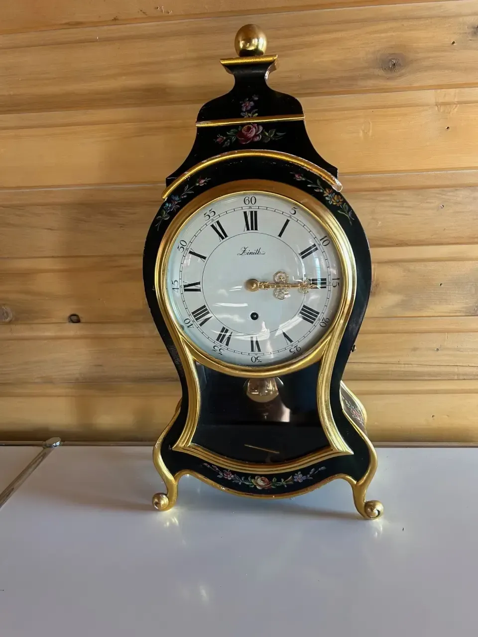 A black and gold clock is sitting on a table in front of a wooden wall.