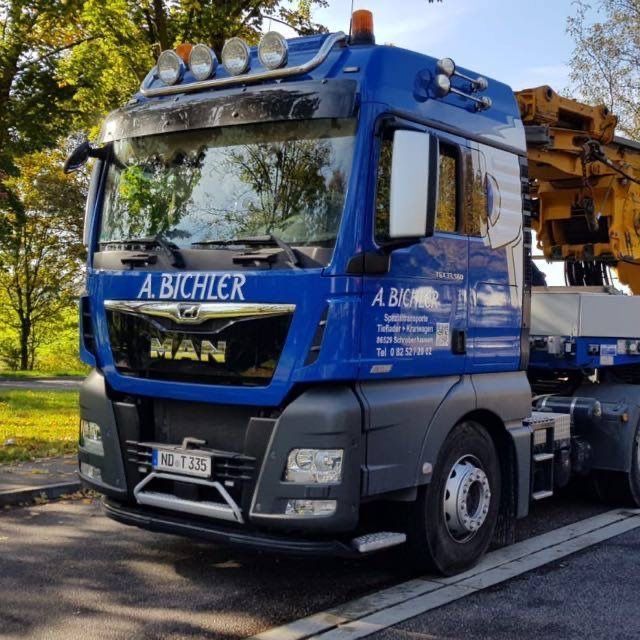 Ein blauer MAN-Baufahrzeug mit der Aufschrift „A. BICHLER“ parkte an einem sonnigen Tag im Freien neben Bäumen.