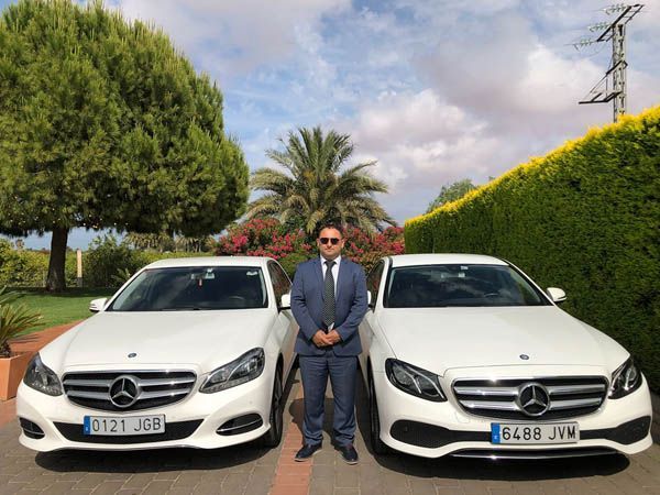 Un hombre con traje y corbata está de pie junto a dos coches Mercedes Benz blancos.