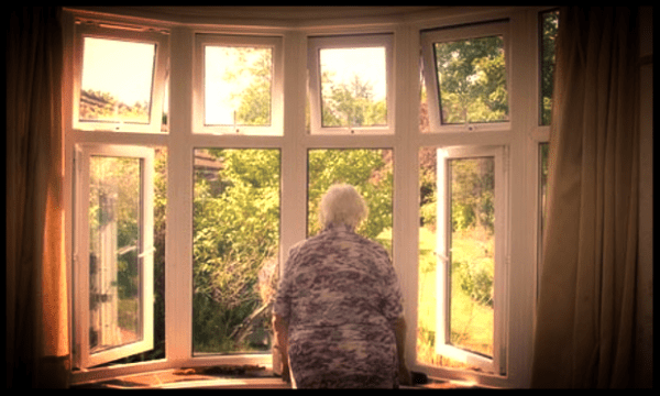 Vanhempi henkilö katsoo avoimesta erkkeri-ikkunasta aurinkoista vihreää puutarhaa.
