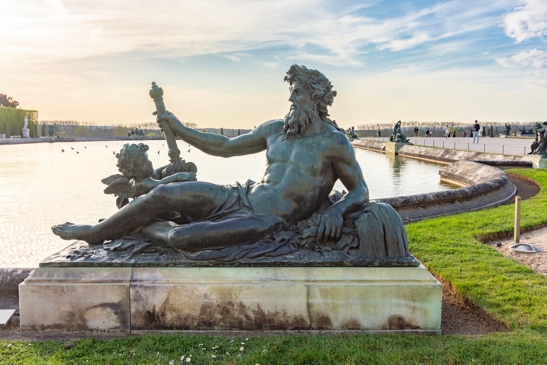 Statue en bronze d'une déesse fluviale allongée, au coucher du soleil, près d'une promenade sinueuse au bord de l'eau.