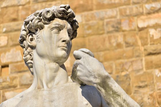 Statue en marbre d'un homme aux cheveux bouclés devant un mur de pierre baigné de soleil.