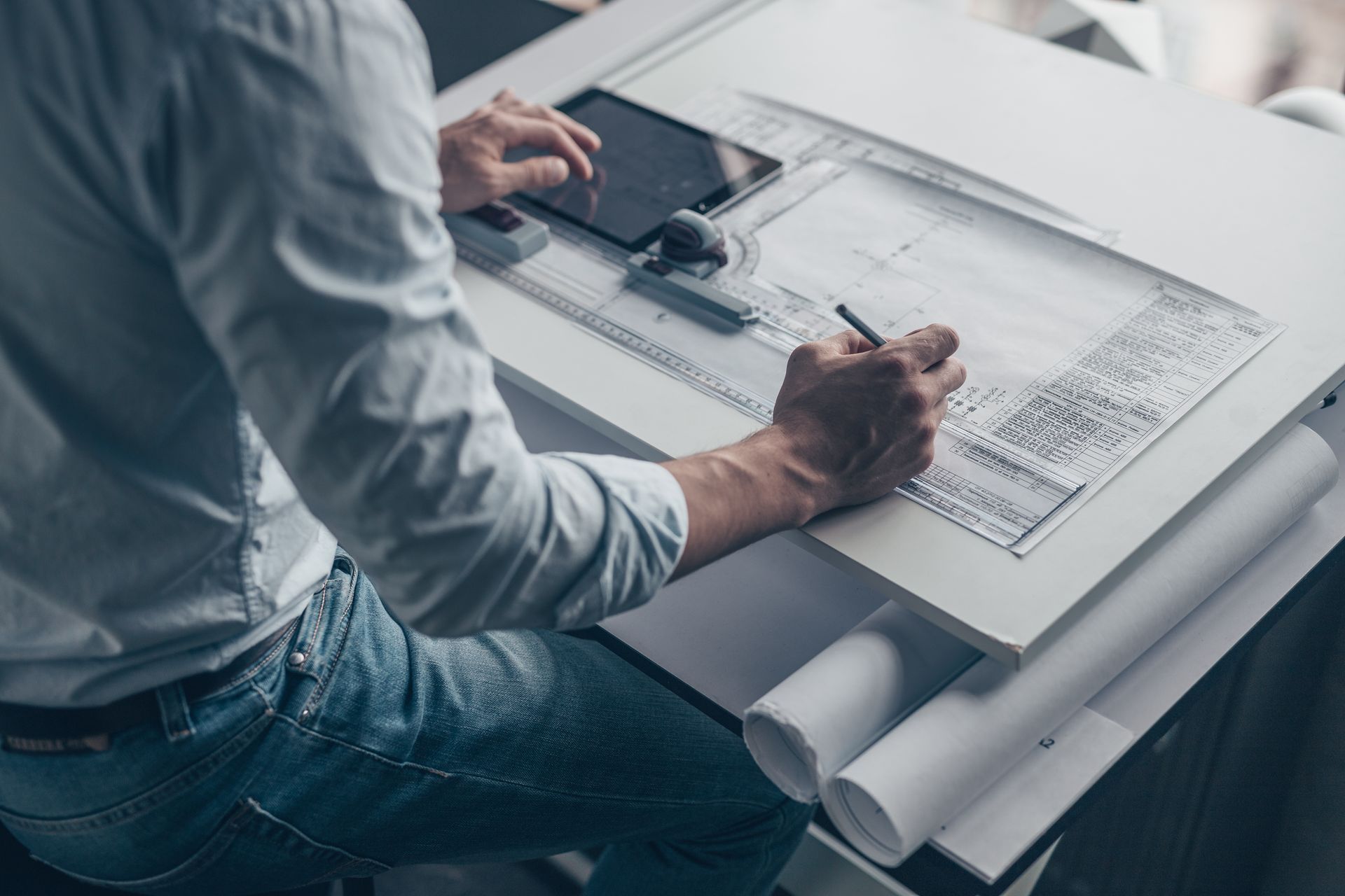 Une personne dessine le design d'un produit sur une grande table à dessin, à l'aide de plans et d'un ordinateur portable.