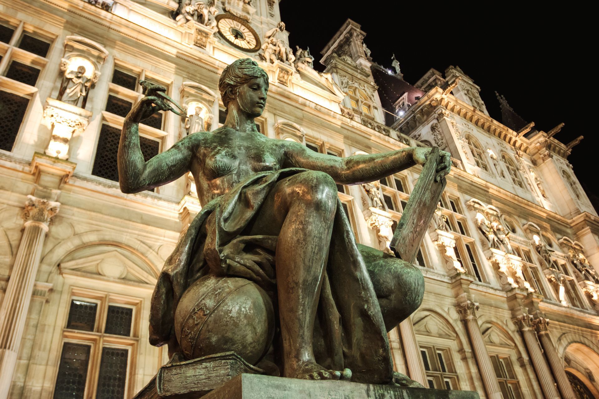 Statue en bronze assise devant un bâtiment historique orné et illuminé la nuit.