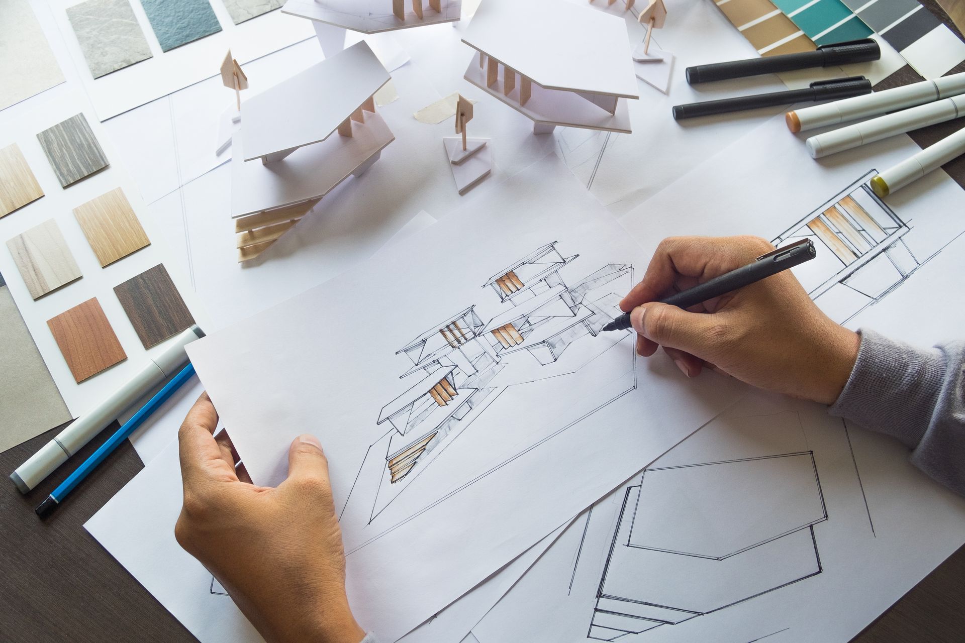 Des mains dessinent le plan d'une maison avec des stylos, des échantillons de couleurs et des maquettes architecturales sur un bureau.